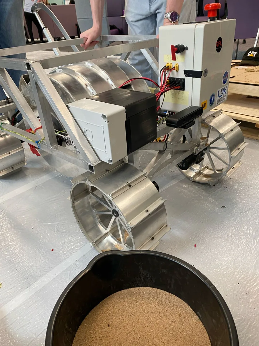 A rover beside a bucket of sand used as regolith